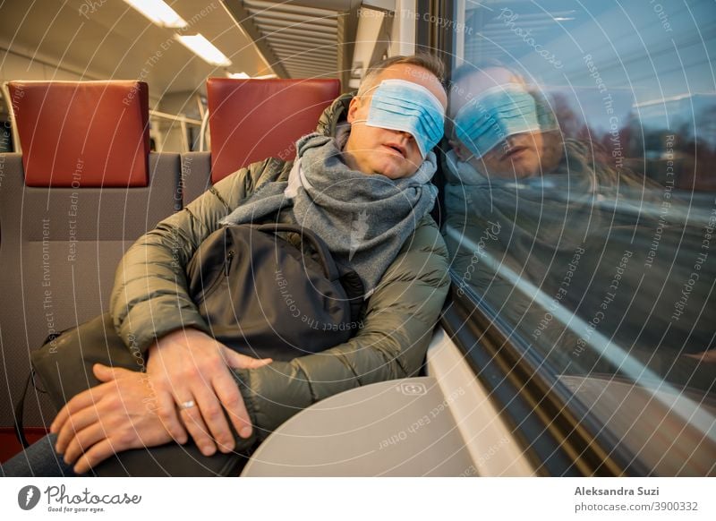 Reisen in öffentlichen Verkehrsmitteln während der Pandemie. Ein müder Mann mittleren Alters mit einer medizinischen Schutzmaske auf dem Gesicht schläft im Nahverkehrszug.