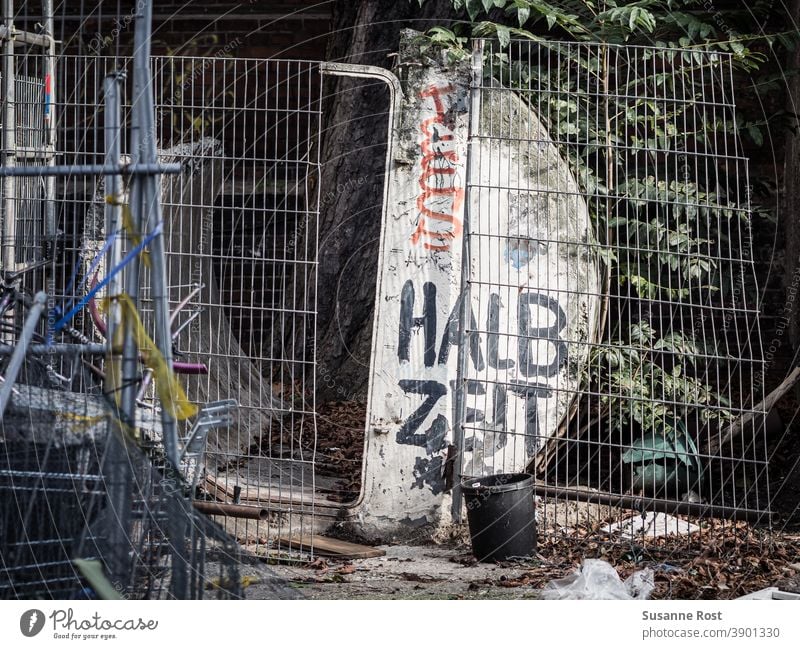 Ein kaputter Bauzaun, davor und dahinter Bauschutt, mit Graffiti bemalt. Mit schwarzer Farbe ist das Wort Halbzeit auf ein halbrundes Bauelement geschrieben.