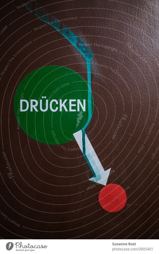 grüner Aufkleber mit dem Hinweis "Drücken", darunter ein weißer Pfeil, der auf einen roten Punkt zeigt. Der Hintergrund ist braun. Graffiti hinweisen