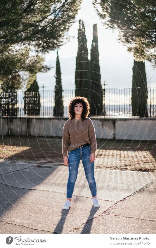 Schöne Frau mit lockigem Haar in einem Park trägt einen beigen Pullover und Jeans mit weißen Turnschuhen. Sonnenlicht Mode Porträt Afro-Look echte Frau Mensch