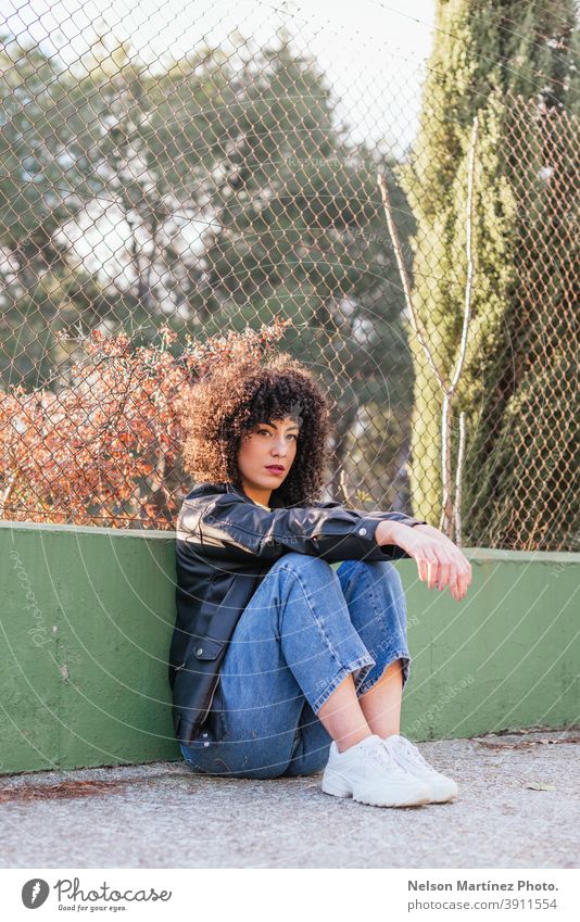 Schöne Frau mit lockigem Haar in einem Park, die eine Lederjacke und weiße Turnschuhe trägt. Sonnenlicht Mode Porträt Afro-Look echte Frau Mensch Ethnizität