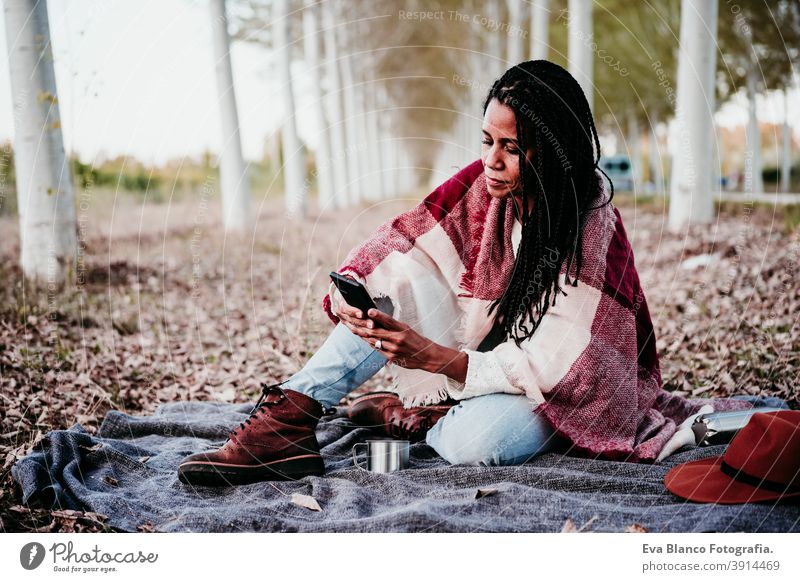 Porträt von hispanischen Mitte erwachsene Frau im Freien halten in Decke eingewickelt. Mit Handy während Picknick.Herbstsaison Afrofrau Latein Sonnenuntergang