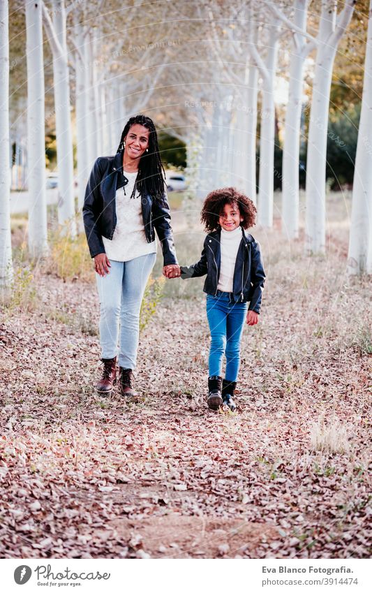 hispanische Mutter und afro Kind Mädchen im Freien zu Fuß bei Sonnenuntergang durch den Weg der Bäume. Herbst-Saison. Familie und Liebe Konzept laufen Umarmung