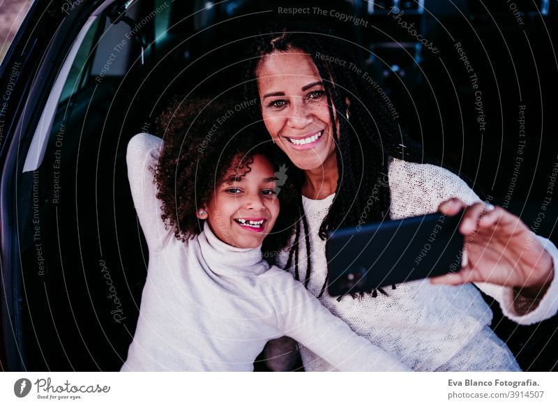 hispanische Mutter und afro Kind Mädchen sitzen in einem Auto in der Natur. Sie haben Spaß und machen Fotos mit dem Mobiltelefon. Herbst-Saison. Familie und Reisen Konzept