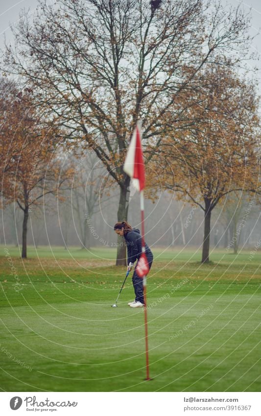 Fitte Golferin mittleren Alters beim Einlochen auf dem Grün von der Fahne aus gesehen, konzentriert auf den nächsten Schlag Frau Winter Herbst windig neblig