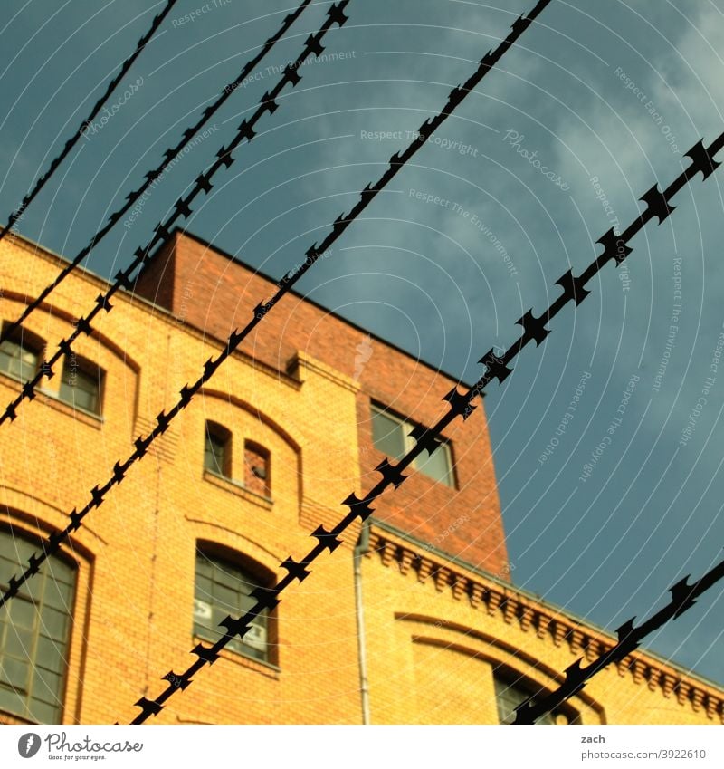 hinter Gittern Haus Stacheldraht Stacheldrahtzaun Backsteinfassade Backsteinhaus gelb diagonal Linie Zaun gefangen Schutz Sicherheit