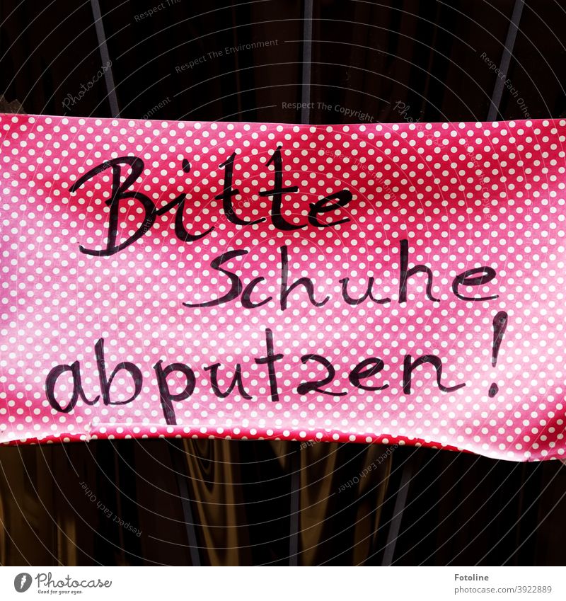 "Bitte Schuhe abputzen" steht handgeschrieben auf einem pinken Schild mit weißen Punkten, welches an eine Glastür geklebt ist. Hinweisschild