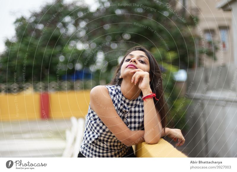 Porträt einer schönen und jungen indischen Bengali brunette Mädchen in westlichen Kleid lehnt auf der Seite der Dachterrasse am Nachmittag mit einem Baum voller gelber Blumen im Hintergrund. Indischer Lebensstil