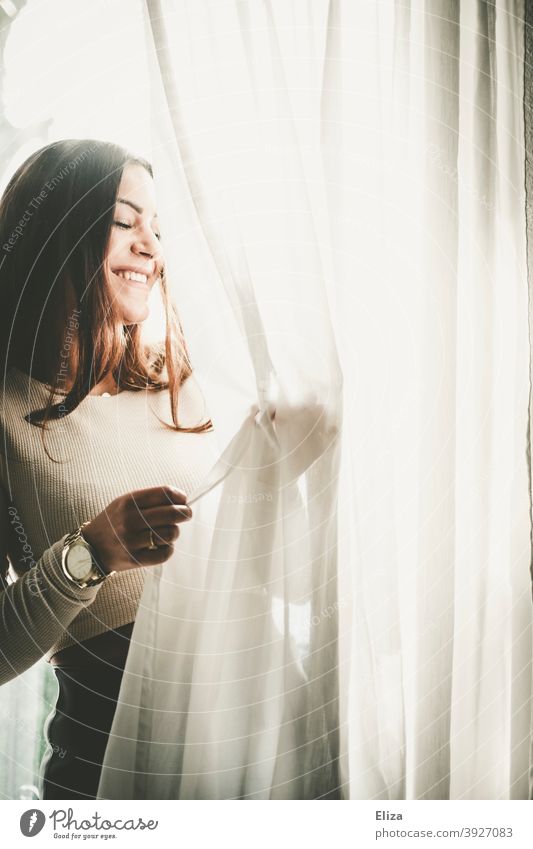 Lächelnde Frau steht am Fenster im Gegenlicht und spielt mit dem Vorhang lächelnd Freude lachen gute Laune Sonne sonnig hell Fröhlichkeit Junge Frau Porträt