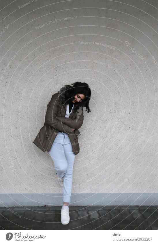 Frau, sich an eine Wand anlehnend frau wand stehen nachdenklich angelehnt jacke jeans schwarzhaarig langhaarig sneakers mauer lächeln glücklich