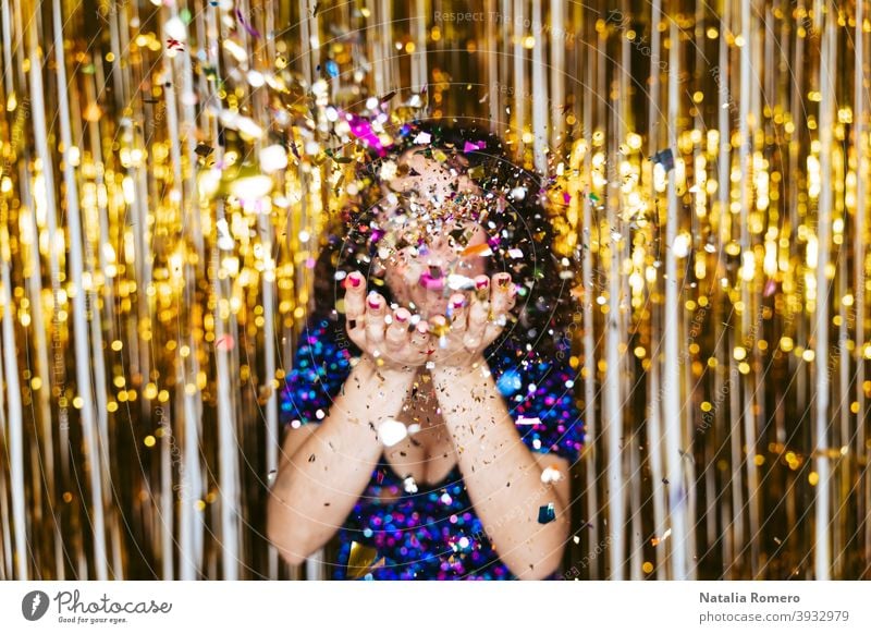 Selektiver Fokus. Eine Frau in einem eleganten glänzenden Kleid und Weihnachtsdekorationen hinter ihr bläst etwas Konfetti in die Kamera. Silvester Party Konzept