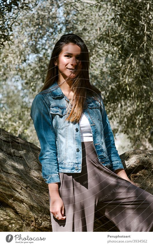 Schöne langhaarige Teenager-Frau mit braunen Haaren lehnt an einem Baum Lehnen grün Natur brünett Gras Kofferraum lässig Landschaft Jeansstoff Grasland