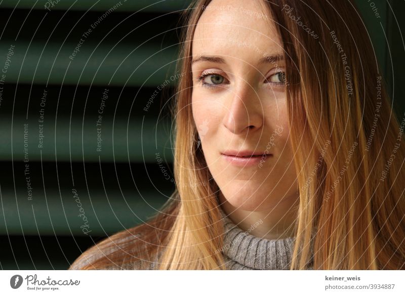 Portrait einer Frau mit schmalem Gesicht vor grüner Jalousie Pullover grüner Hintergrund Kopf weiblich schmunzeln Lippen Nase blond Mund Haare & Frisuren Auge