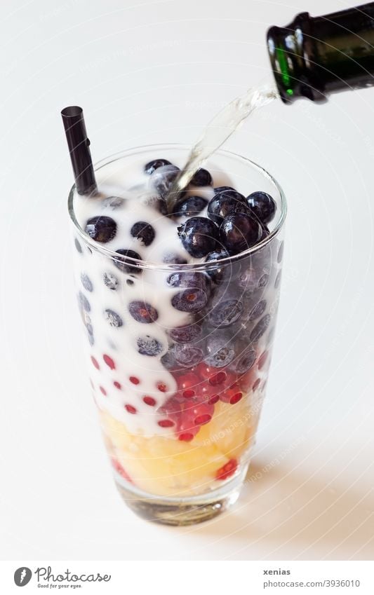 In ein Glas mit schwarzem Trinkhalm, Blaubeeren, roten Johannisbeeren und gelben Ananas wird schäumender Sekt geschüttet - Prost Party Sektflasche Sylvester