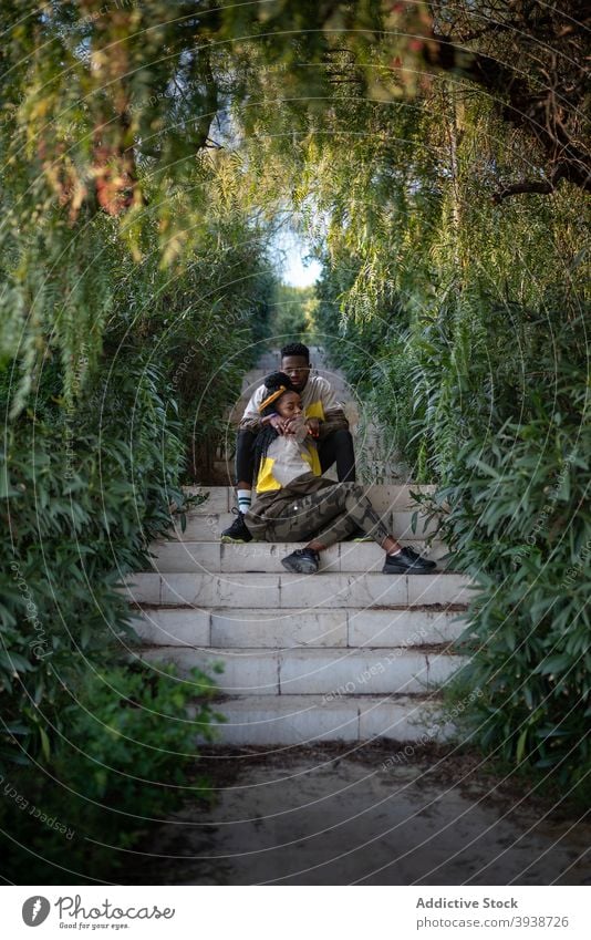 Junge trendige schwarze ethnische Paar umarmt auf der Treppe im grünen Park Umarmung Garten Liebe romantisch Zusammensein Zuneigung Baum Natur Umarmen