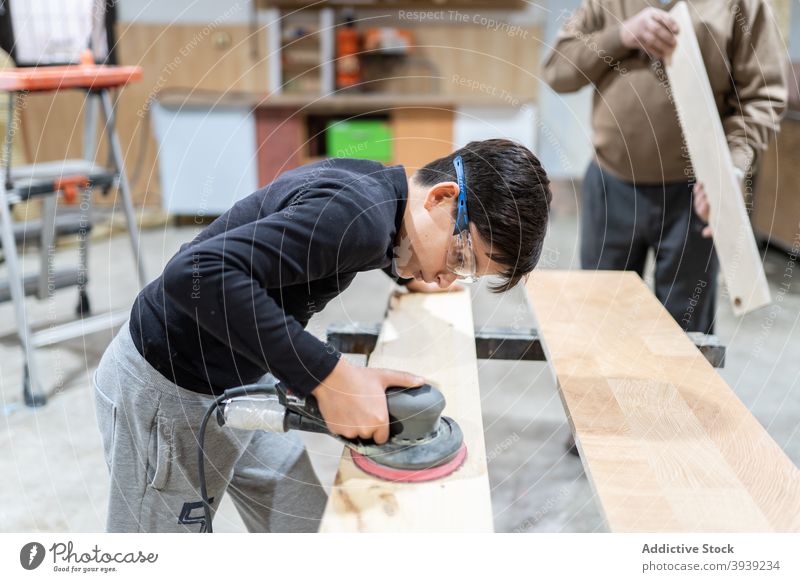 Konzentriertes Kind beim Polieren von Holz in der Werkstatt mit unkenntlichem Großvater Junge polnisch Schleifmaschine Maschine Hilfsbereitschaft Enkel Orbital