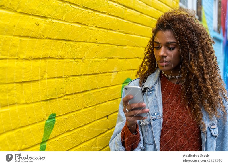 Selbstbewusste junge schwarze Frau mit Handy und zu Fuß entlang der Straße in der Nähe von mehrfarbigen Gebäude Spaziergang Smartphone Graffiti farbenfroh