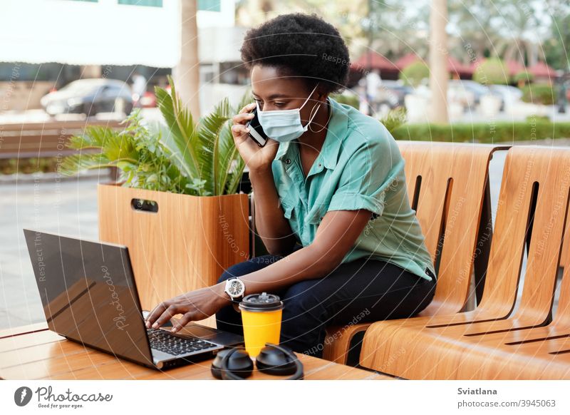 Ein junges afrikanisches Mädchen mit einer medizinischen Gesichtsmaske telefoniert und arbeitet mit einem Laptop an einem Tisch in einem Café. Soziale Distanzierung und Arbeit, Arbeit online, Business online