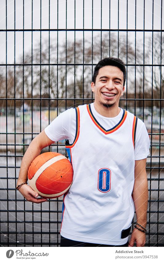 Latino Kerl posiert mit einem Basketball durch den Zaun von einem Gericht im Freien. Sport-Konzept. Lachen freundlich Spieler Individualität lachen Motivation