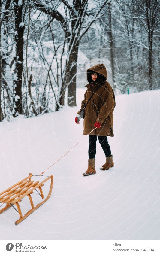 Eine Frau mit einem Schlitten im Schnee im Winter kalt verschneit Natur Wald Mantel braun Kapuze Holzschlitten Wintertag winterlich