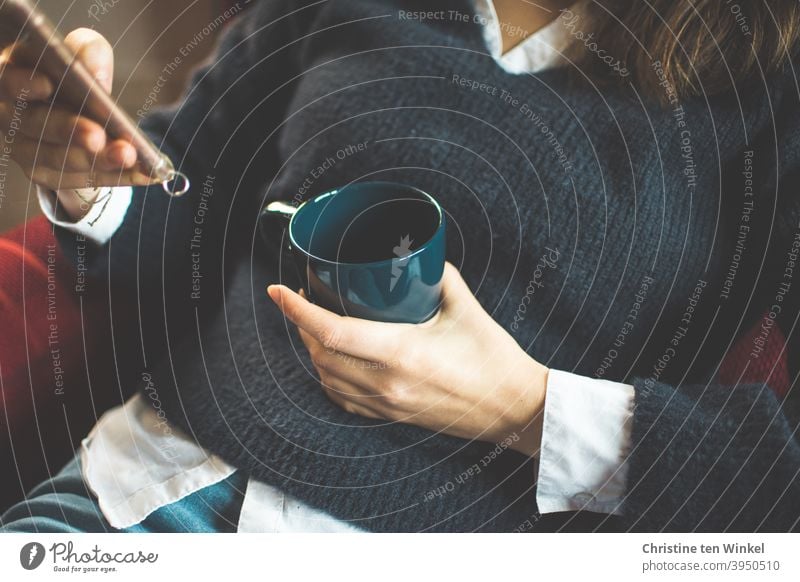 Die junge Frau im blauen Pullover sitzt gemütlich im Sessel und hält Kaffeebecher und Smartphone in ihren Händen. Oberkörper Portrait ohne Kopf Hand festhalten