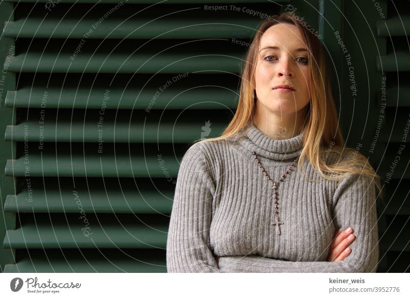 Eine Frau mit schmalem Gesicht trägt Rosenkranz um den Hals und steht angelehnt an grüner Jalousie Pullover grüner Hintergrund Kopf Portrait Religion weiblich