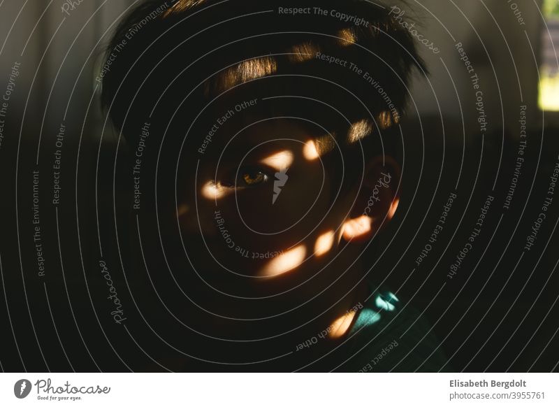 Kleiner Junge, mit Blick in die Kamera und Lichteinfall der Sonne auf das Auge, was durch die kleinen Öffnungen der Rollläden hindurchgeht Lichteinfall Auge