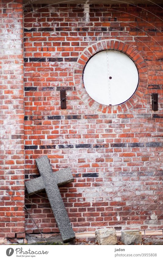 christliches kreuz aus stein lehnt schräg an einer mauer, welche eine runde öffnung hat. Religion & Glaube Kreuz Christliches Kreuz Christentum Kirche