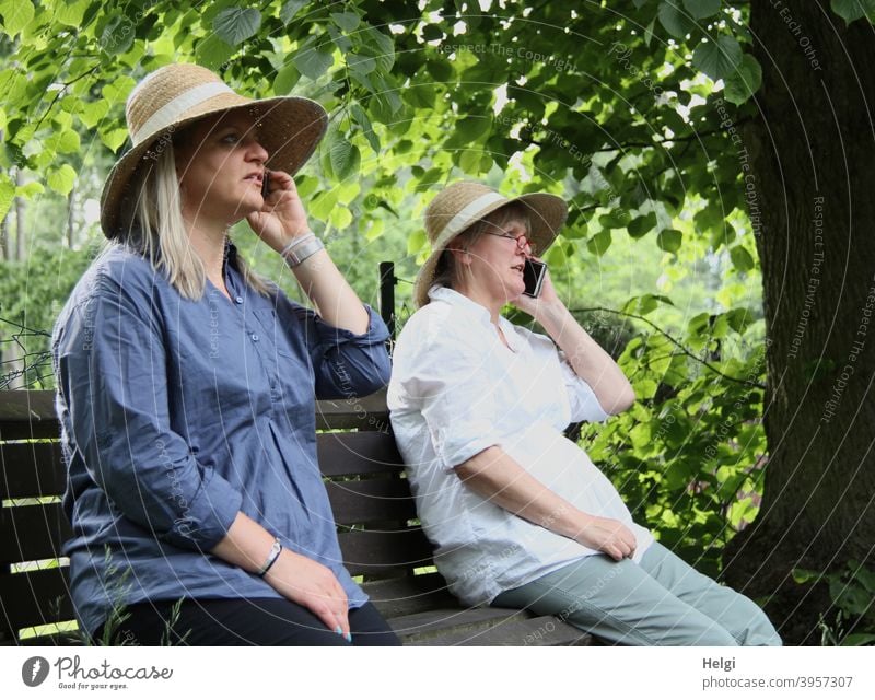 "Wurde wirklich Zeit,dass wir uns wiedersehen! Ich freu mich!" - Zwei Frauen mit Hut sitzen auf einer Bank und telefonieren miteinander Mensch Seniorin Baum