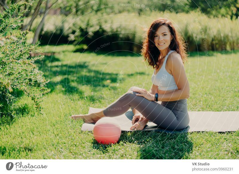 Positive brunette Frau in abgeschnittenen Top und Leggings posiert nackten Füße auf karemat verwendet Ball für gymnastische Übungen lächelt angenehm genießt schöne Natur Blick. Sport im Freien. Yoga-Praxis.