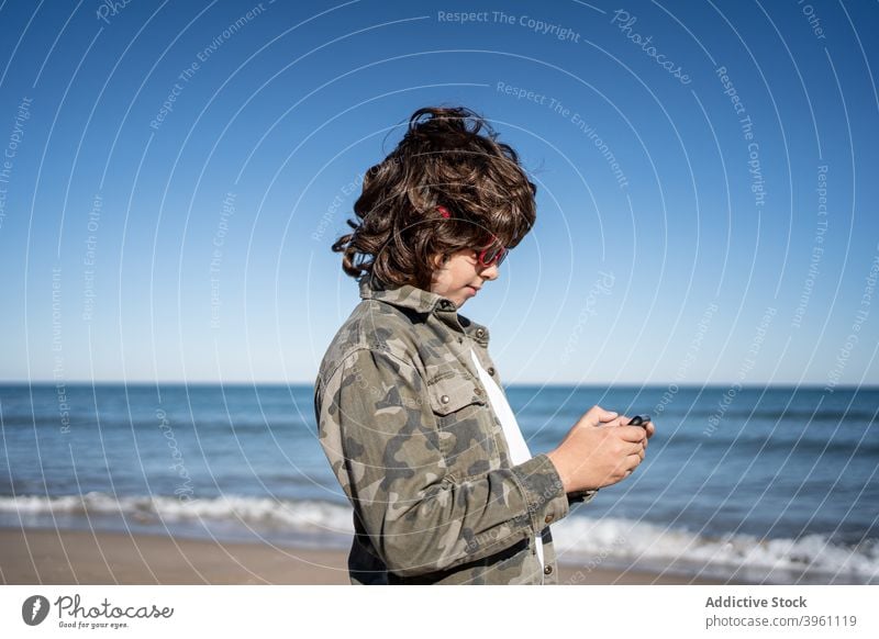 Junge mit Sonnenbrille am Strand spielen mit einem Handy Smartphone authentisch aktiv Pause Kind Kinder Küste Konzept Spaß Feiertag Lifestyle Natur im Freien