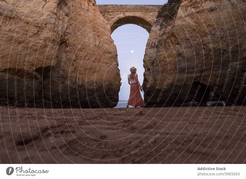 Anonyme Dame zu Fuß auf sandigen Meeresufer in der Nähe von gewölbten Klippen unter Sonnenuntergang Himmel Frau Spaziergang Strand Bogen Abend Feiertag