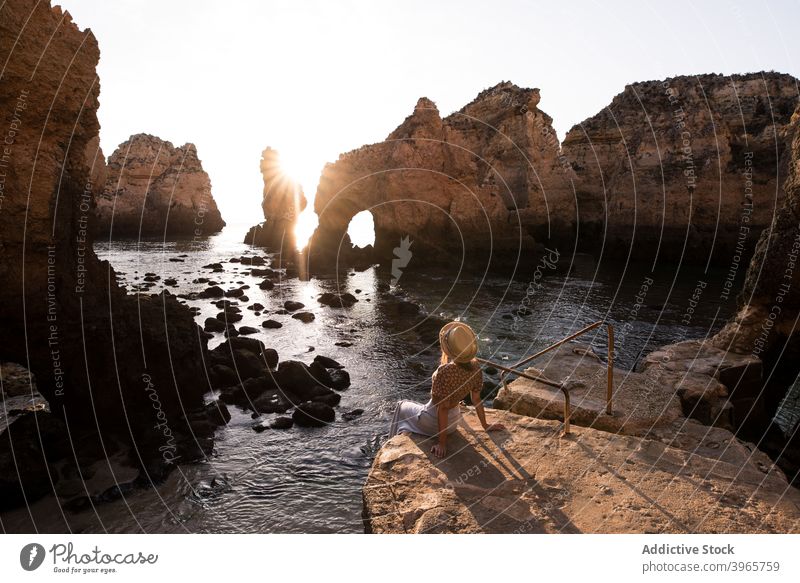 Unerkennbare Frau rastet in der Nähe von Meer und Klippen ruhen MEER bewundern Sonnenuntergang Wasser Natur Küste Stein Algarve Portugal Urlaub sitzen Hut