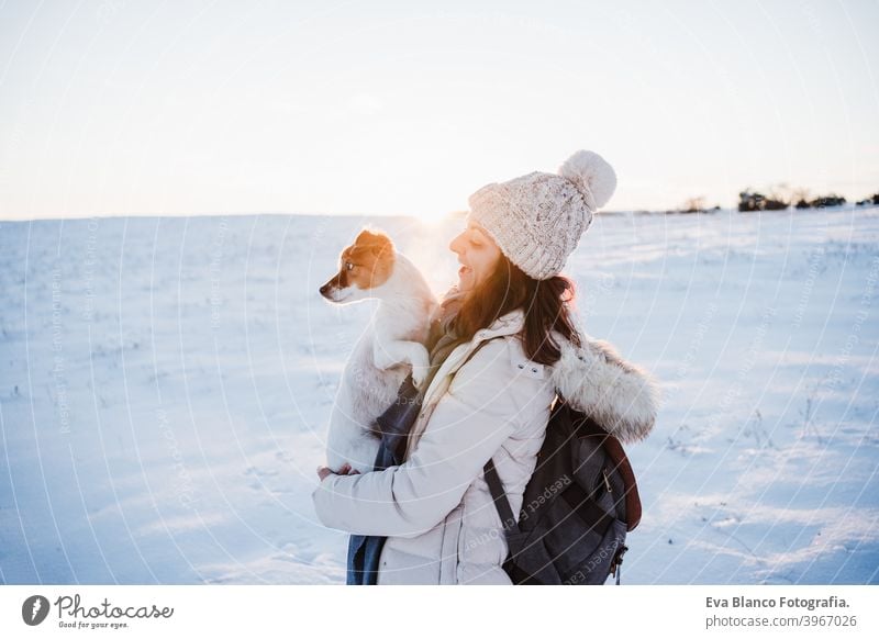 glückliche Frau in verschneiten Berg trägt modernen Mantel bei Sonnenuntergang. Halten niedlichen Jack Russell Hund in den Armen. Wintersaison. Natur bei Sonnenuntergang