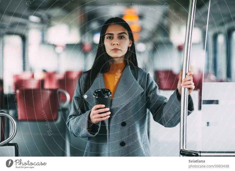 Eine fröhliche Brünette mit Kaffee in der Hand steht im Zug, schaut in die Ferne und lächelt. Verkehr jung Frau schön Eisenbahn Passagier U-Bahn Lächeln