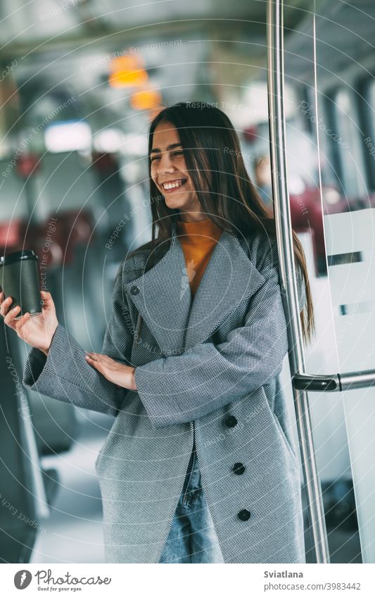 Eine fröhliche Brünette mit Kaffee in der Hand steht im Zug, schaut in die Ferne und lächelt. Verkehr jung Frau schön Eisenbahn Passagier U-Bahn Lächeln