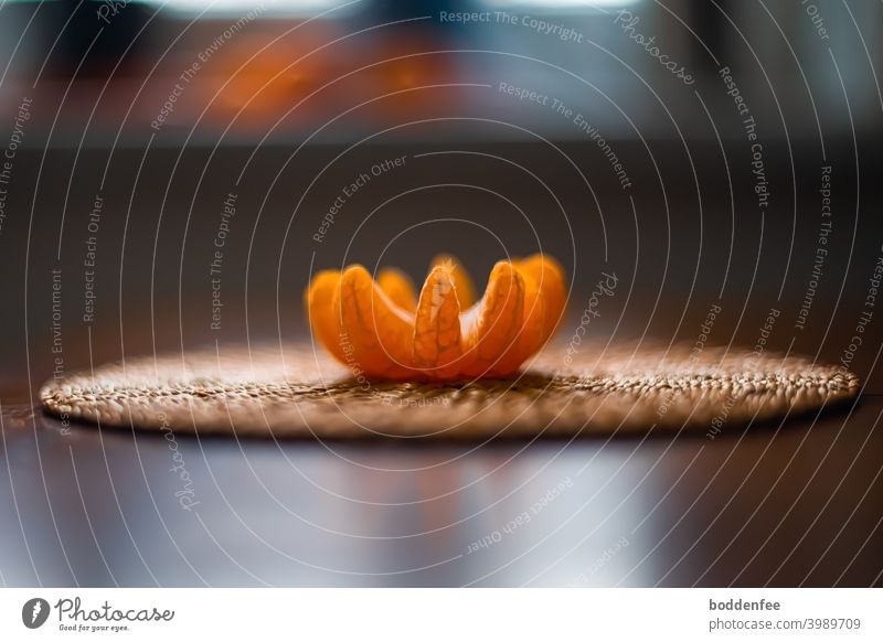 Eine geschälte und aufgefaltete Clementine liegt auf einem Untersetzer aus Reisstroh. Aus der Höhe der Tischplatte im Gegenlicht fotografiert, fokussiert auf die Clementine