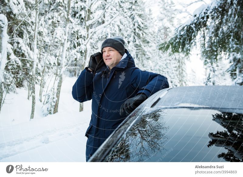 Mann in warmer Winterkleidung steht am Auto, mit Telefon. Snowy Winter Landstraße, Auto mit Eis bedeckt, Schöne Wald unter dem Schnee. Hilfsbereitschaft Anruf