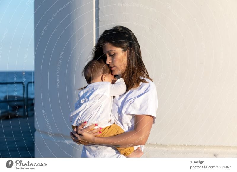 Junge Mutter hält ihr kleines Baby gegen weiße Wand in sonnigen Tag Kind neugeboren Liebe Familie Frau Pflege Beteiligung wenig Mutterschaft jung Kindheit