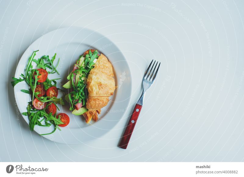 gesundes leckeres Frühstück - Croissant mit Avocado, Rucola, Schinken und Käse serviert mit Kirschtomatensalat auf weißem Teller Belegtes Brot Lebensmittel