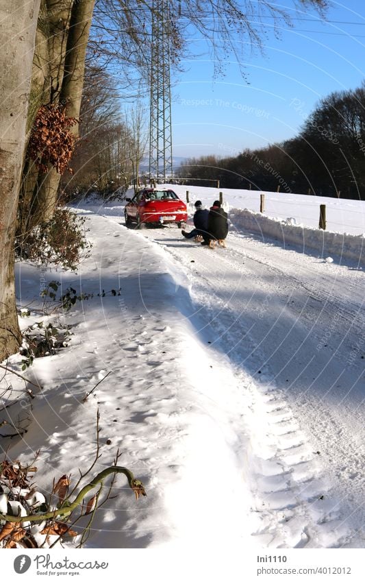 rotes Auto zieht Schlitten mit zwei Personen durch schneebedeckte Winterlandschaft Winterfreude Winterfreuden verschneite Straße verschneite Landschaft