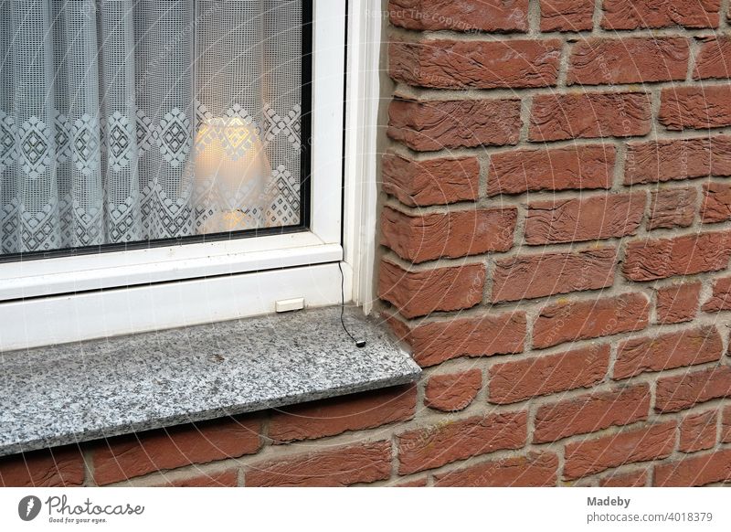 Schlichte Tischleuchte hinter einer spießigen Gardine in einem Fenster mit weißem Rahmen in rotbrauner Backsteinfassade in Oerlinghausen im Teutoburger Wald in Ostwestfalen-Lippe