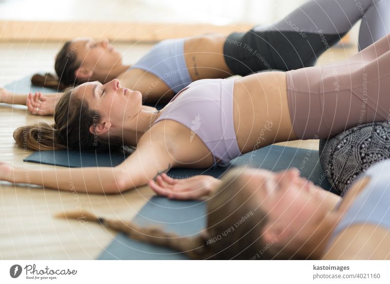 Restorative Yoga mit einem Bolster. Gruppe von drei jungen, sportlichen, attraktiven Frauen im Yogastudio, liegend auf einem Bolster-Kissen, dehnend und entspannend während Restorative Yoga. Gesunder aktiver Lebensstil