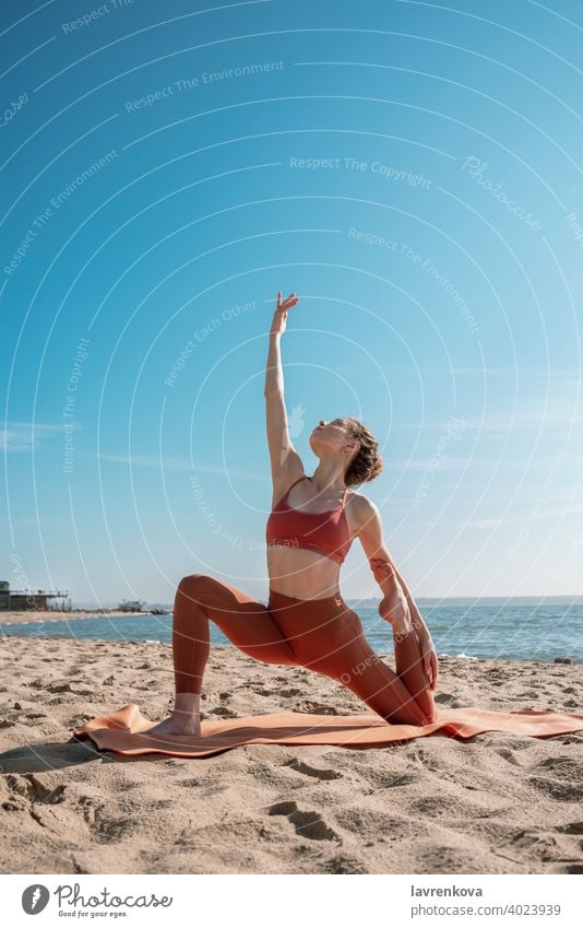 Junge Frau in orangefarbenem Oberteil und Leggins beim Üben der Mondsichel-Pose (Anjaneyasana-Variante) an einem Strand am Morgen Übung Yoga jung praktizieren