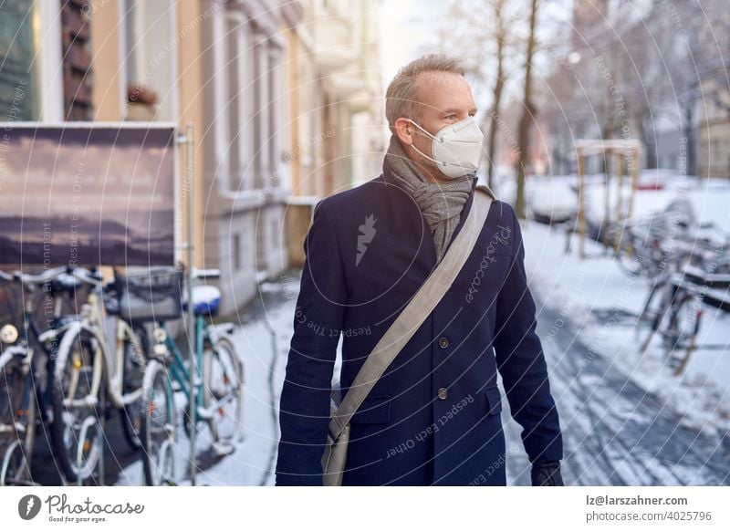 Mann mit einer schützenden chirurgischen Gesichtsmaske während der Covid-19- oder Coronavirus-Pandemie und Wintermantel mit Ledertasche über der Schulter, der eine verschneite städtische Straße mit geparkten Fahrrädern in Nahaufnahme entlanggeht und zur Seite schaut