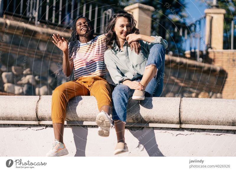 Zwei Freunde, die sich gemeinsam auf der Straße amüsieren und auf einer Stadtmauer sitzen. Frauen multiethnisch schwarz Afro-Look Mädchen Schüler zwei Spaß