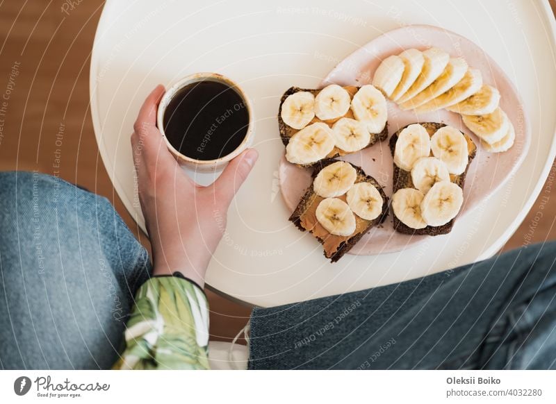 Frau hält eine Tasse schwarzen Kaffee, Schuss direkt über. Mit gesundem Obst und Erdnussbutter-Sandwich zum Frühstück, trinken Morgen Kaffee zu Hause Banane