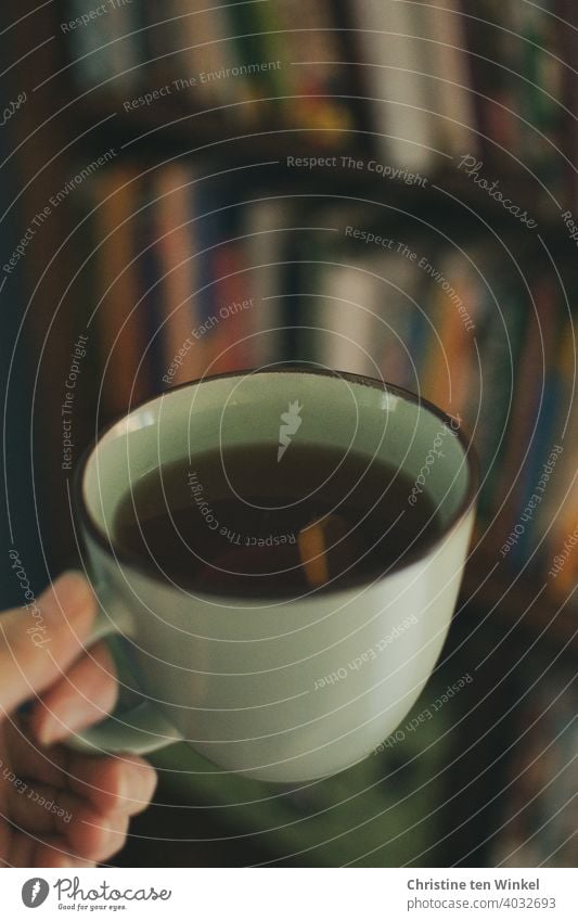 Teetasse mit schwarzem Tee und Zitrone in einer Hand, dahinter unscharf ein Bücherregal Darjeeling Winter Teebecher aufwärmen kälte Frühstück heiß Pause