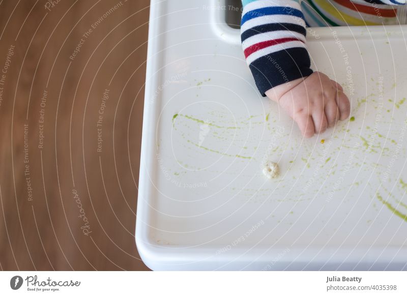 Junges Baby in Regenbogenstreifen-Outfit sitzt am Hochstuhl und greift nach Puffmüsli mit grünem Püree auf dem Tablett; babygeführte Entwöhnung Zange palmar