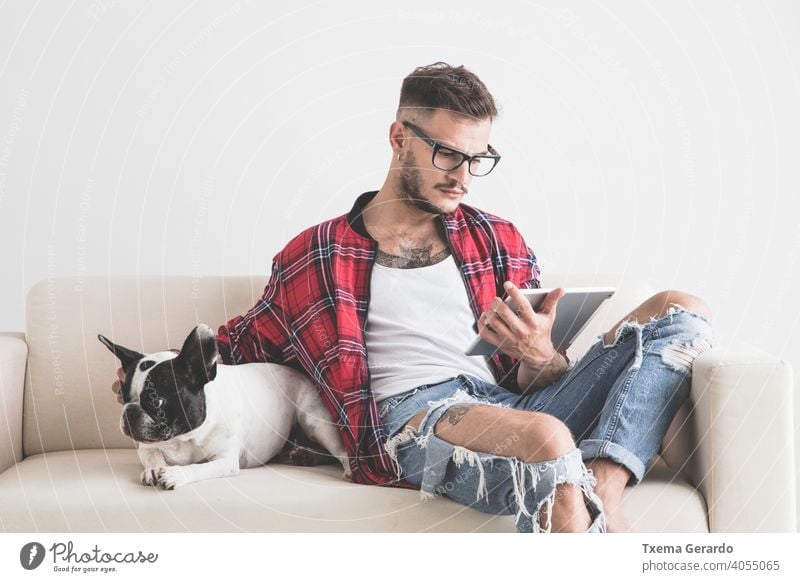 Junge Hipster Blick auf sein Tablet neben seinem Hund sitzt auf dem Sofa französische Bulldogge Piercing durchbohrt Hut Ohrringe Hemd Vollbart Haustier Welpe